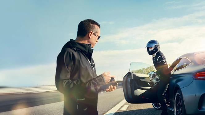 Ein Mann mit einem Handy grüßt einen Fahrer mit Helm, der ein AMG Fahrzeug verlässt