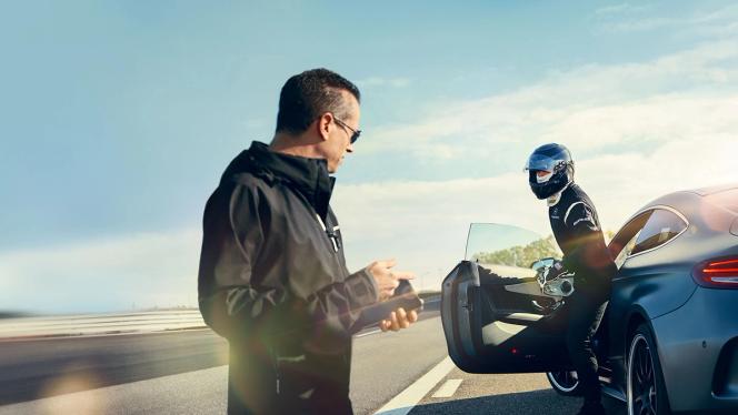 Ein Mann mit einem Handy grüßt einen Fahrer mit Helm, der ein AMG Fahrzeug verlässt