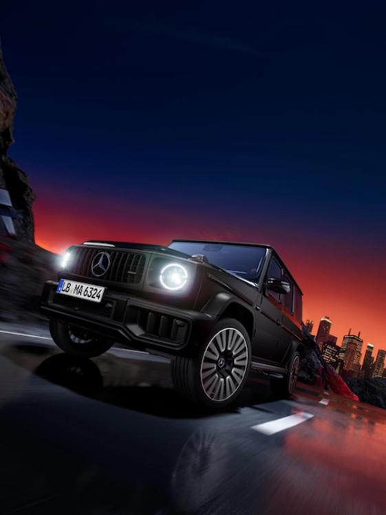 Mercedes-AMG G63 mit Two-Finish Paket in schräger Vorderansicht mit leuchtenden Scheinwerfern auf schwarzer Fläche mit Felsen und Sonnenuntergang im Hintergrund
